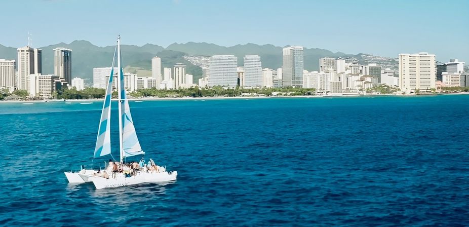 Catamaran with blue-and-white sails carrying passengers on deep-blue ocean off Waikiki, Honolulu skyline and green mountains along the shoreline under a clear sky