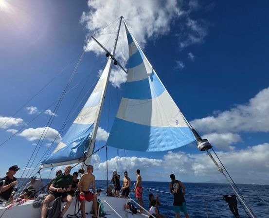 Sunlit sailing trip on the open ocean: group of people in swimwear aboard a blue-and-white sailboat with a large striped sail against a bright blue sky with fluffy clouds.
