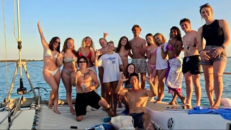 Group of smiling young adults in swimsuits posing on a sailboat at golden-hour sunset over calm coastal waters, carefree summer boat-party vibe