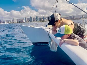 Adult and child leaning over the rail of a sailboat, peering into deep blue ocean with a distant coastal city skyline under a bright, partly cloudy sky.