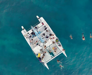 Aerial view of a white catamaran with passengers and swimmers in clear turquoise tropical waters, people snorkeling and relaxing on deck during a boat tour.