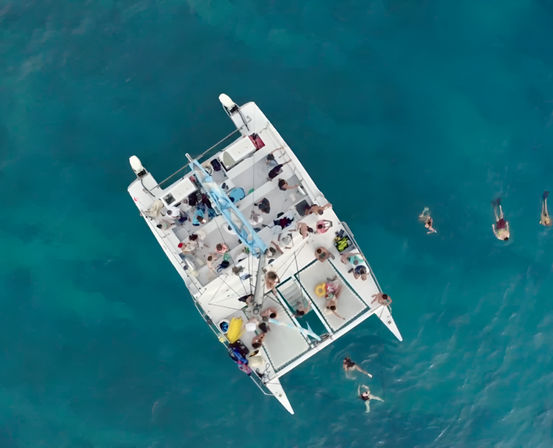 Aerial view of a white catamaran with passengers and swimmers in clear turquoise tropical waters, people snorkeling and relaxing on deck during a boat tour.