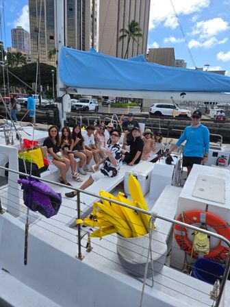 Group of people relaxing on a white catamaran at an urban marina with a blue sail cover, palm trees and high-rise buildings in the background, yellow kayaks and an orange life ring on deck
