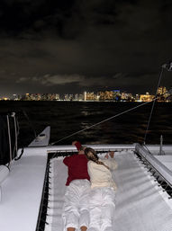 Two people lying side-by-side on a catamaran trampoline during a cozy night sail, looking toward an illuminated urban skyline across dark water under a cloudy sky.