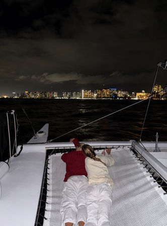 Two people lying side-by-side on a catamaran trampoline during a cozy night sail, looking toward an illuminated urban skyline across dark water under a cloudy sky.
