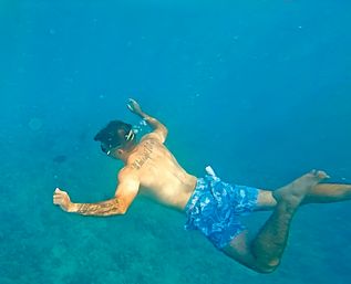 Person snorkeling in clear blue tropical ocean, wearing a mask and blue floral swim trunks, gliding over a shallow seabed with tattoos on their back and arm and bubbles trailing.