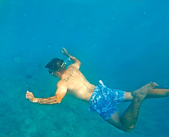 Person snorkeling in clear blue tropical ocean, wearing a mask and blue floral swim trunks, gliding over a shallow seabed with tattoos on their back and arm and bubbles trailing.