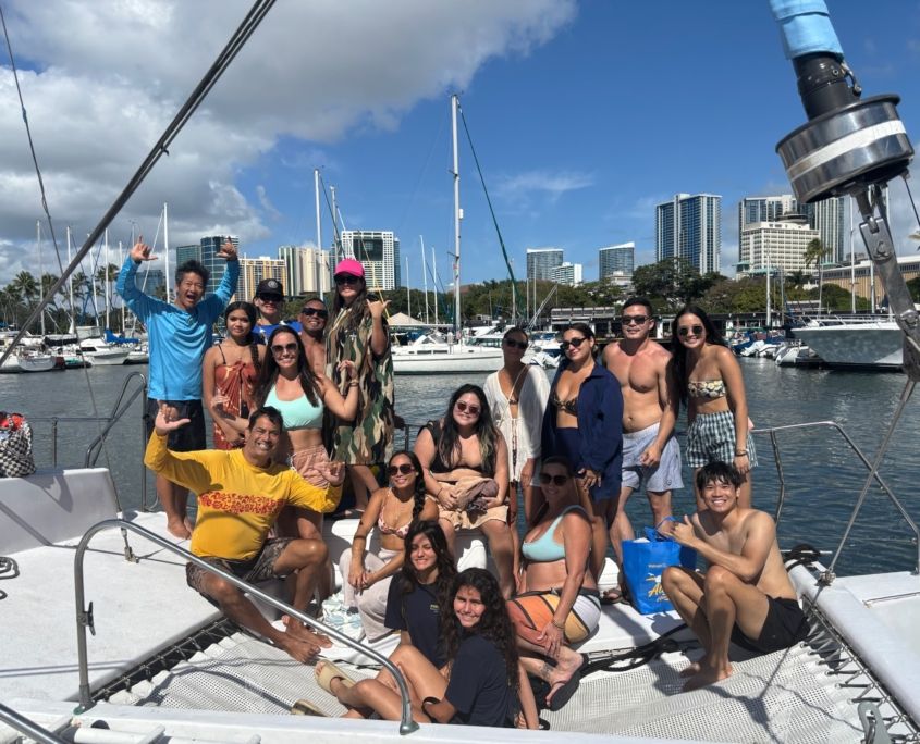 Lively group of friends in swimsuits posing and making shaka signs on a sailboat at a sunny marina, with moored yachts, palm trees and a coastal city skyline under blue skies.