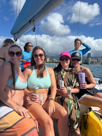 Four women smiling on a sunny sailboat off Waikiki, Honolulu, with city skyline, mountains, blue sky and drinks in hand.