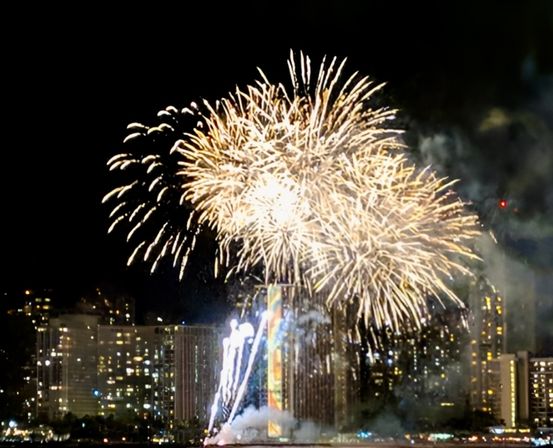 Golden fireworks bursting over a nighttime waterfront city skyline, high-rise buildings lit up with lights and reflections on the water.