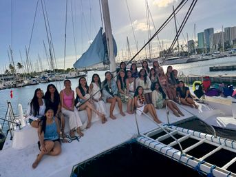 Cheerful group of about 20 young women seated on the deck of a white catamaran at a sunny marina, surrounded by sailboats, palm trees and waterfront high-rise buildings.