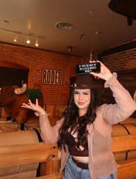 Smiling woman in a cowgirl hat posing next to a mechanical bull inside a western-themed bar with exposed brick and a framed RODEO sign, holding up a phone sign.