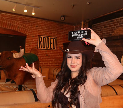 Smiling woman in a cowgirl hat posing next to a mechanical bull inside a western-themed bar with exposed brick and a framed RODEO sign, holding up a phone sign.
