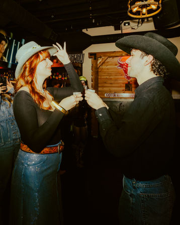 Two people wearing cowboy hats clinking shot glasses in a lively western-themed bar, woman tipping her hat and man smiling with rustic wood decor and neon bull sign in the background.