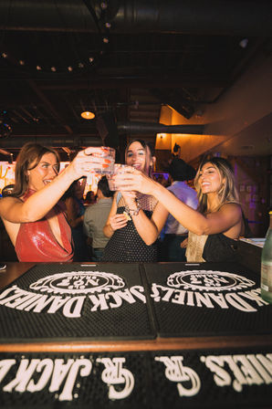 Three women clinking drinks over a bar counter in a lively, dimly lit urban bar — smiling and toasting on a festive night out.