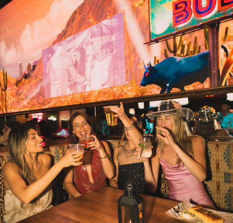 Four women toasting drinks at a lively cowboy-themed bar with neon signage, desert mural and bull display, one wearing a sparkling cowboy hat — girls' night out, urban nightlife.