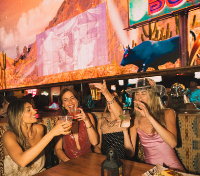 Four women toasting drinks at a lively cowboy-themed bar with neon signage, desert mural and bull display, one wearing a sparkling cowboy hat — girls' night out, urban nightlife.