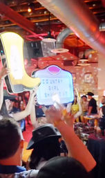 Lively country bar scene: crowd in cowboy hats holding a neon yellow boot and a light-up marquee reading 'COUNTRY GIRL SHAKE IT' under disco lights and exposed ductwork.