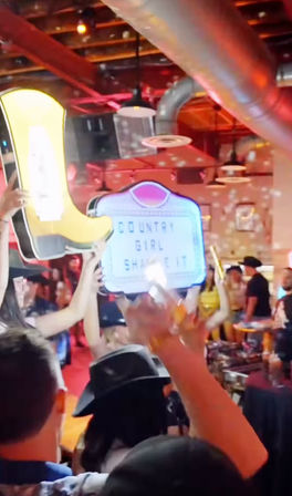 Lively country bar scene: crowd in cowboy hats holding a neon yellow boot and a light-up marquee reading 'COUNTRY GIRL SHAKE IT' under disco lights and exposed ductwork.