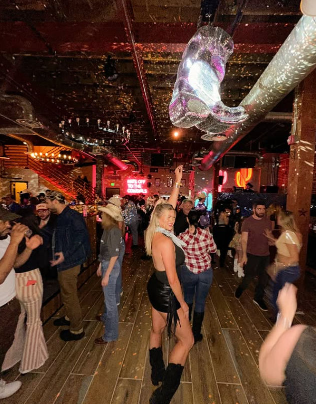 Woman in a black mini dress and boots dancing with one arm raised on a crowded wooden dance floor at a lively rustic bar, neon signs, disco lights, confetti and partygoers in cowboy hats under exposed ceiling pipes.