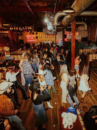 Crowded country bar dance floor with people in cowboy hats and boots line dancing under rustic wooden beams and warm string lights