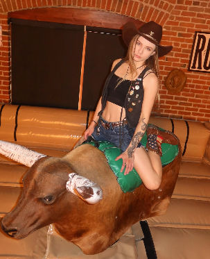 Person in a cowboy hat, black crop top and denim shorts riding a mechanical bull with a green saddle in a western-style indoor space with exposed brick walls and padded arena.