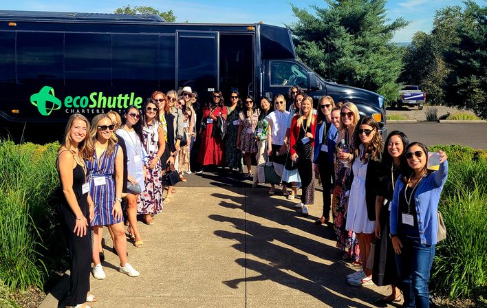 Smiling group of event attendees wearing badges lined up along a sunny walkway beside a black shuttle bus with green branding and landscaped greenery in the background.