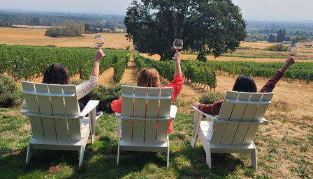 Three people in white Adirondack chairs toasting with wine glasses while overlooking neat vineyard rows, a large oak tree and rolling countryside on a sunny wine-tasting afternoon.
