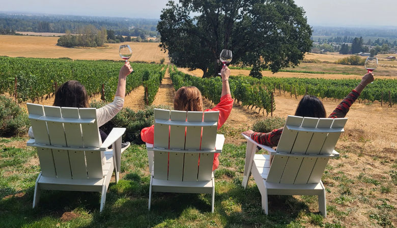 Three people in white Adirondack chairs toasting with wine glasses while overlooking neat vineyard rows, a large oak tree and rolling countryside on a sunny wine-tasting afternoon.