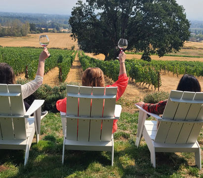 Three people in white Adirondack chairs toasting with wine glasses while overlooking neat vineyard rows, a large oak tree and rolling countryside on a sunny wine-tasting afternoon.