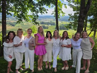 Group of ten women in light summer dresses smiling and holding wine glasses beneath trees with a vineyard and rolling countryside view