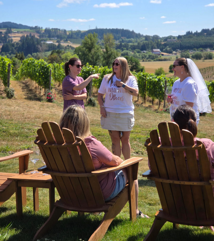Women enjoying a summer wine tasting at a vineyard: a bride-to-be wearing a veil holds a glass while friends chat by wooden Adirondack chairs with grapevines and rolling hills behind them.
