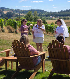 Women enjoying a summer wine tasting at a vineyard: a bride-to-be wearing a veil holds a glass while friends chat by wooden Adirondack chairs with grapevines and rolling hills behind them.
