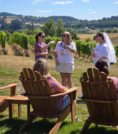 Women enjoying a summer wine tasting at a vineyard: a bride-to-be wearing a veil holds a glass while friends chat by wooden Adirondack chairs with grapevines and rolling hills behind them.