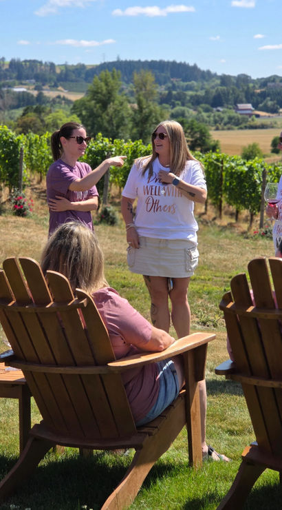 Women enjoying a summer wine tasting at a vineyard: a bride-to-be wearing a veil holds a glass while friends chat by wooden Adirondack chairs with grapevines and rolling hills behind them.