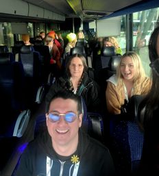 Cheerful group of travelers on a coach bus interior — selfie of a man wearing reflective glasses in the foreground with seated passengers behind, bright window light