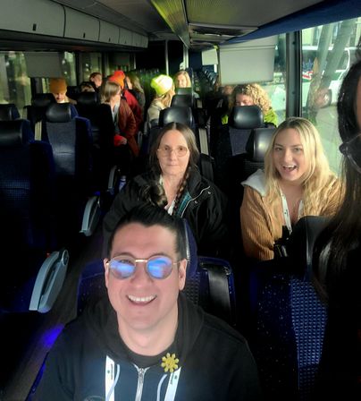 Cheerful group of travelers on a coach bus interior — selfie of a man wearing reflective glasses in the foreground with seated passengers behind, bright window light