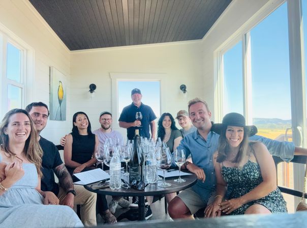 Smiling group of nine enjoying a wine tasting in a bright winery tasting room, wine glasses and bottles on a table with a panoramic vineyard and countryside view through large windows.