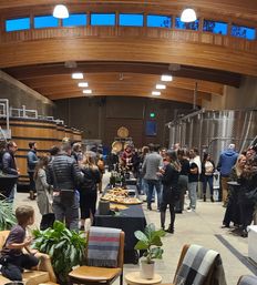 Winery tasting event in a vaulted wood‑ceiling room with oak barrels and stainless steel fermentation tanks, guests mingling around tables with wine bottles and charcuterie, lounge chairs and potted plants in the foreground.