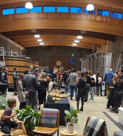 Winery tasting event in a vaulted wood‑ceiling room with oak barrels and stainless steel fermentation tanks, guests mingling around tables with wine bottles and charcuterie, lounge chairs and potted plants in the foreground.