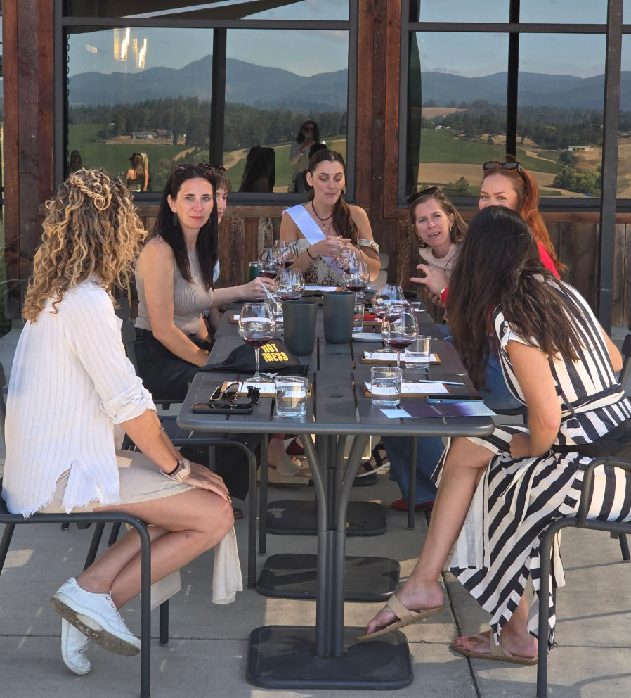 Group of women enjoying a wine tasting on a sunny winery patio, wine glasses on a long table and rolling vineyard hills reflected in the windows