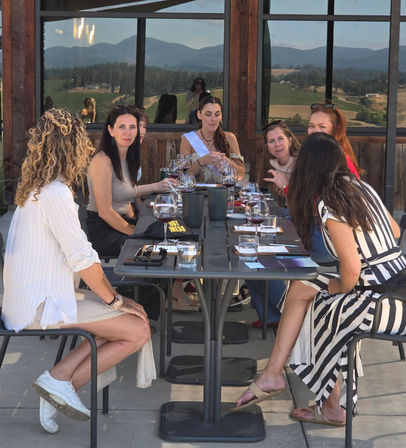 Group of women enjoying a wine tasting on a sunny winery patio, wine glasses on a long table and rolling vineyard hills reflected in the windows