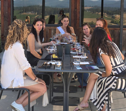 Group of women enjoying a wine tasting on a sunny winery patio, wine glasses on a long table and rolling vineyard hills reflected in the windows