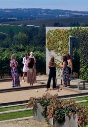 Group of women socializing on a bocce court at a sunny vineyard, surrounded by grapevines and rolling green hills — casual summer dresses, hats and pastel bocce balls.
