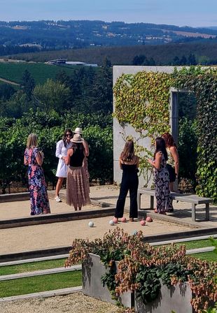 Group of women socializing on a bocce court at a sunny vineyard, surrounded by grapevines and rolling green hills — casual summer dresses, hats and pastel bocce balls.