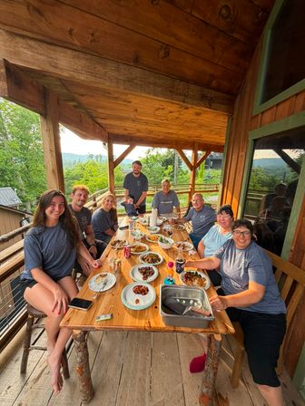 Smiling group sharing a cozy, family-style meal on a rustic wooden mountain cabin deck around a long table of plates and bowls, with lush green hills and trees visible beyond the railing.