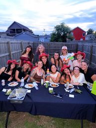 Dozen women laughing at a suburban backyard party at dusk, many wearing red cowboy hats, gathered around a long table with plates, drinks and playful props.