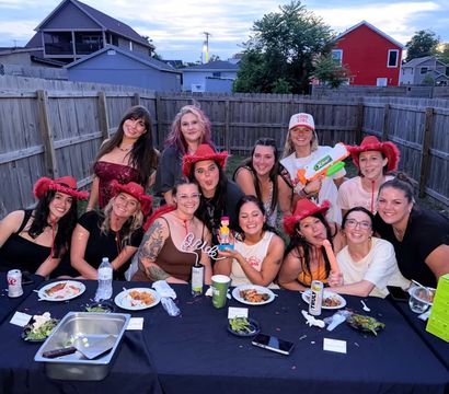Dozen women laughing at a suburban backyard party at dusk, many wearing red cowboy hats, gathered around a long table with plates, drinks and playful props.