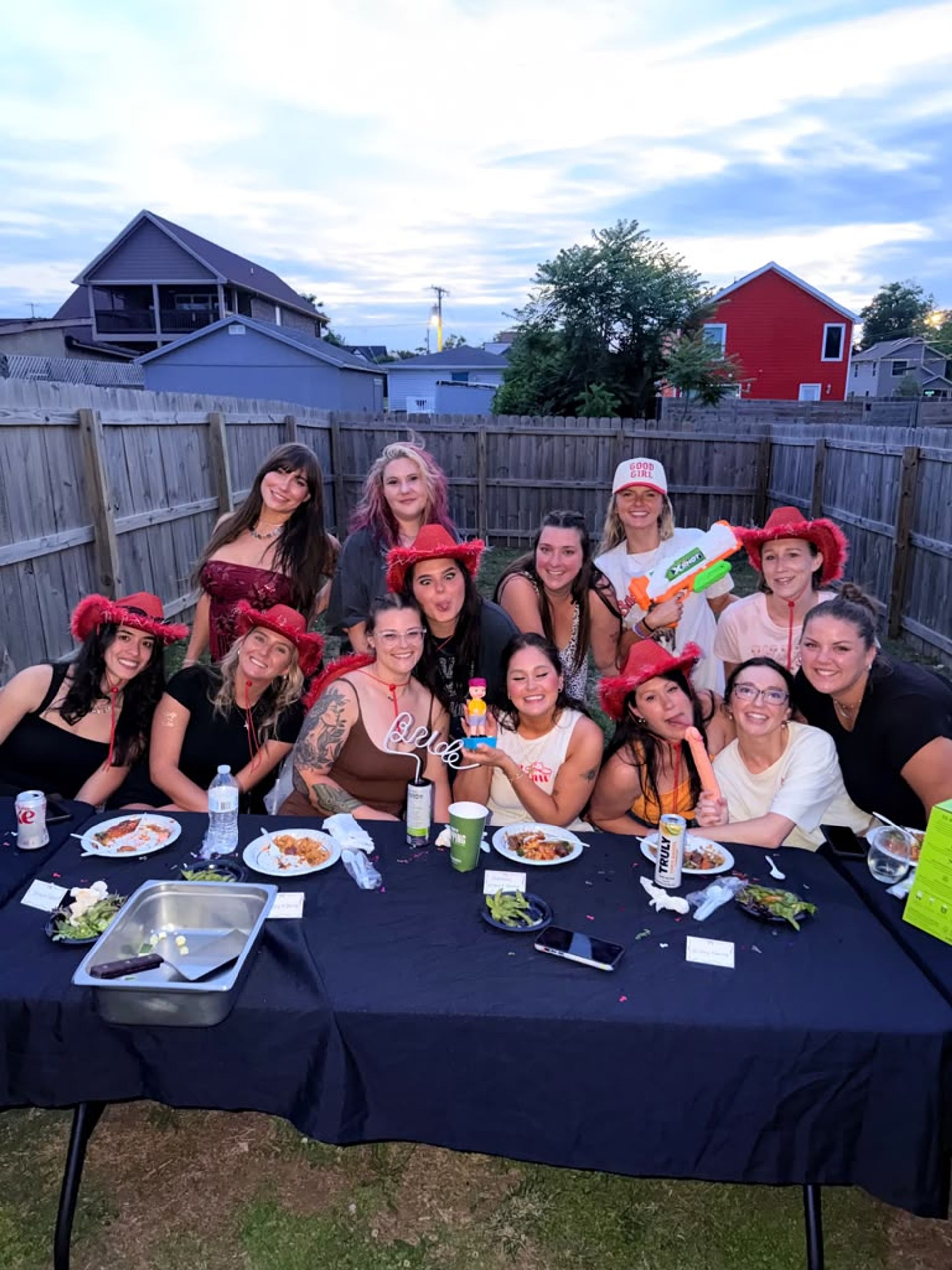 Dozen women laughing at a suburban backyard party at dusk, many wearing red cowboy hats, gathered around a long table with plates, drinks and playful props.