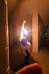 Fire performer tilting back to catch a bright flame in a dim, pillar-lined concrete corridor with warm orange glow and a finger partially in the foreground